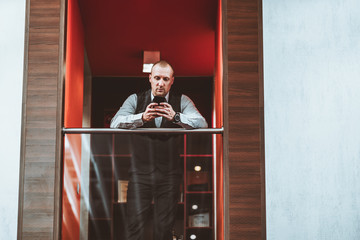 An elegant serious mature man entrepreneur is typing a message via his smartphone while leaning against a chrome railing of a balcony in modern office settings, a copy space place on the right