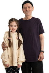 Attractive smiling old man in black hat, t-shirt and white wireless headphones, senior pencioner looking at the camera and hugging his granddaughter dressed in beige hoodie, white wall on the