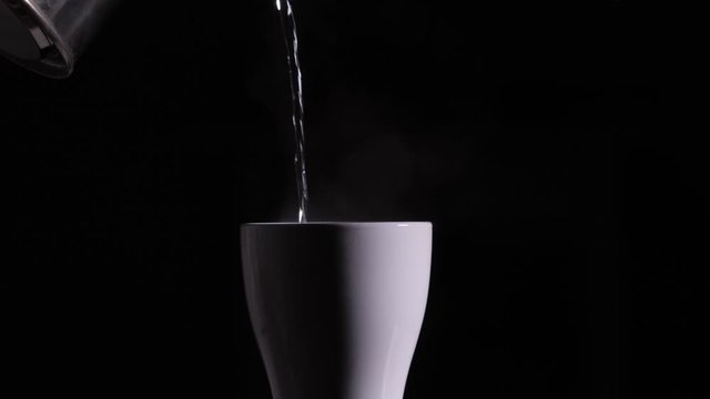 Hot boiled water is poured from a metal kettle into a white ceramic cup or mug on a black background.