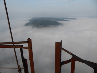 Ocean of fog underneath the top of a smoke stack chimney, hills and mountains rising in the distance, rusted bars and bulbs, good morning