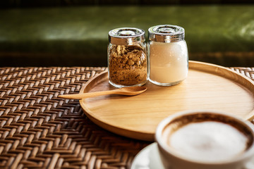 Sugar in pot on wood plate near cup of coffee.