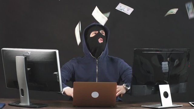 Young anonymous hacker man sit in mask in space full of money, dollar bills fly around the room, incognito dangerous hacker sit isolated over black background