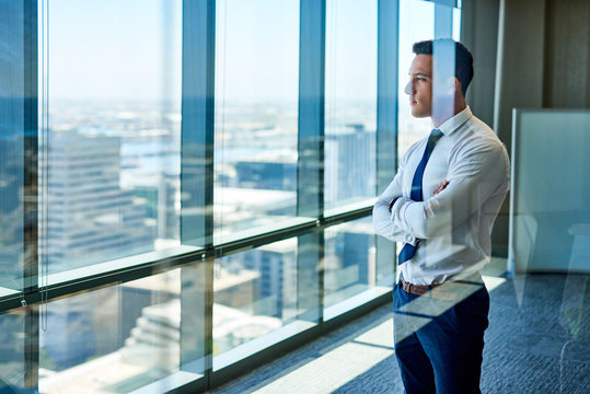 Confident young businessman deep in thought while looking through windows at the city from high up in an office building
