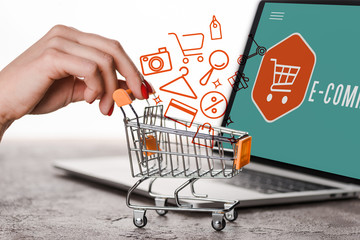 cropped view of woman holding toy shopping cart near laptop and illustration on white, e-commerce...