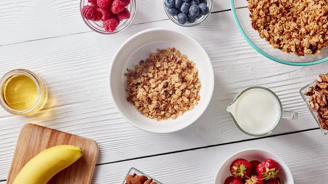 Preparation of healthy breakfast from natural organic products - granola, fruits, berries, walnuts, honey, milk on a wooden white background. Top view. Stop motion animation in 4K