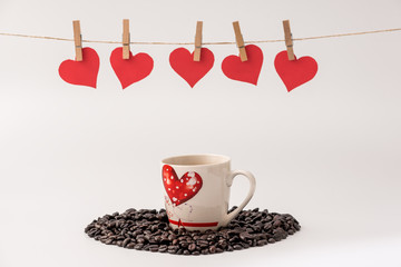 Cup of Coffee with heart made of coffee grains on white background and Clothes pegs and red paper hearts on rope above. Valentines day concept