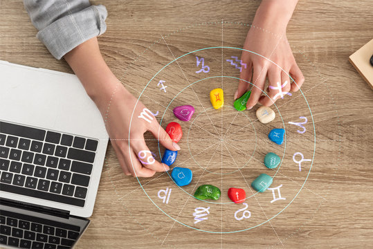Cropped view of woman holding stones with zodiac signs beside laptop and illustration