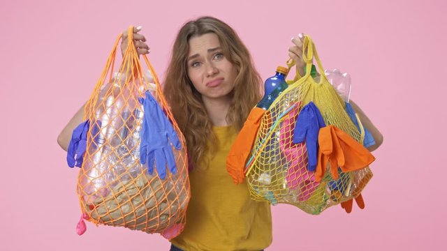 A young sad woman is holding string bags with plastic waste and looking at the camera isolated over pink background