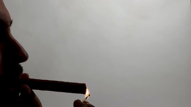 Silhouette of man lightning up the cigar with a lighter and smoking it. Man smoking a cigar. Black smoke on white background.