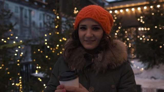 Portrait of an attractive girl in a bright hat with coffee in hands in the winter on a background of lights