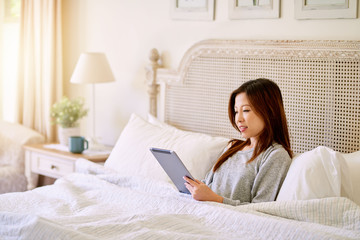 Smiling Asian woman using a tablet while lying in bed