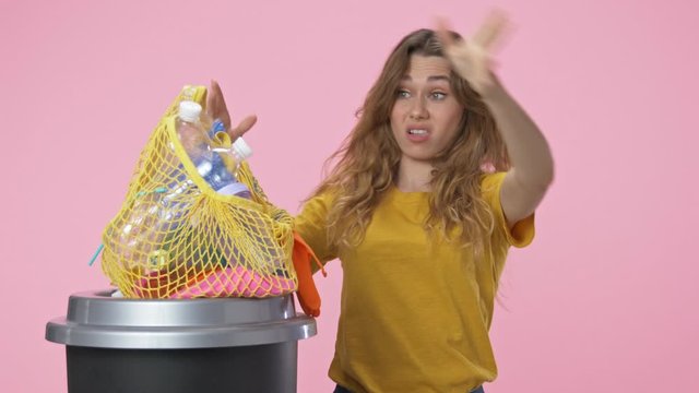 A good-looking young woman starts to sort garbage then puts her string bag to the litter isolated over pink background