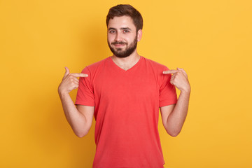 Portrait of handsome bearded young man points at himself, attractive European male standing over yellow background, has pleasant appearance, wearing red casual t shirt. Copy space for advertisment.