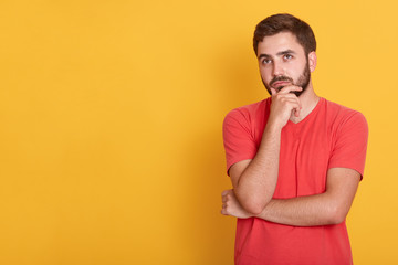 Picture of young cool caucasian man confused, feels doubtful and unsure, keeping hands ubder chin, looking aside, having thoughtful facial expression. Copy space for advertisment or promotional text.