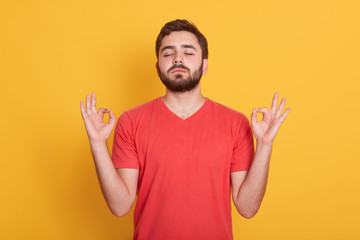 Indoor shot of relaxed carefree bearded male meditates against yellow background, tries to relax after long hours of working, concentrates on something, wering casual clothing., People concept.