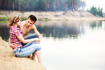 Fototapeta na wymiar Young beautiful loving couple having rest on river shore on summer day with green nature at background. Love, relationships, dating, flirt and attraction concept