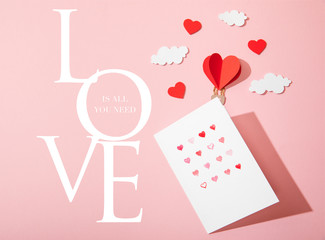 top view of greeting card with hearts near paper heart shaped air balloon in clouds and love is all you need lettering on pink