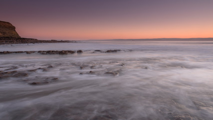 Sea water, washing over a rocky shore, while the sun sets on the horizon. A long exposure has created a milky sea