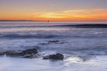 Waves rolling over rocks on the shoreline, while the sun sets over the horizon