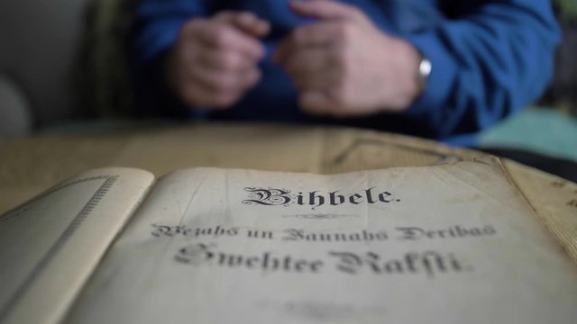 View Of Open Old Latvian Bible On Table Inside Room With Adult Male Wearing Blue Jumper In The Background 