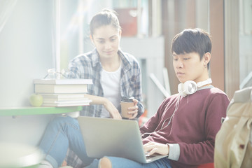 Asian boy sitting and typing on laptop computer while girl sitting near by him and drinking coffee they studying together