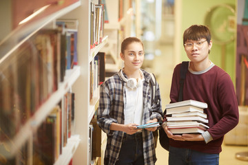 Portrait of two university students with books looking a camera while standing near the bookcase in...