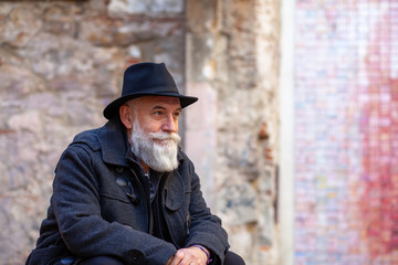 older man with white beard and black hat sitting on a street bench with thoughtful face