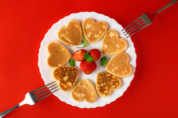 Creative breakfast for Valentine's Day. Heart shaped pancakes with strawberries on a plate with forks, on a red background. Valentine's day background. Top view. Flat lay. Copy space