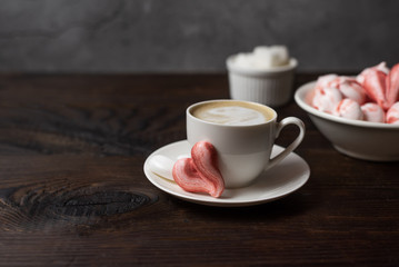 Beautiful romantic breakfast coffee with cream and meringue cookies in the shape of hearts. White cup stand on a dark wooden table with a concrete background. Close up still life with copy space.