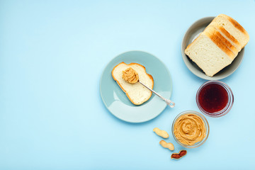 Top view,flat lay process of cooking breakfast, spreading bread, toast with peanut butter, creamy peanut paste on blue colored background. Served by peanut butter, peanuts in shell,jam