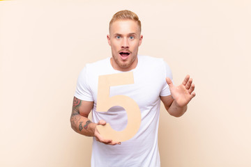 young blonde man surprised, shocked, amazed, holding a number 5.