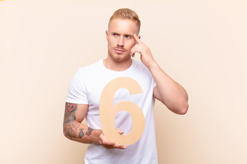 young blonde man confused, doubtful, thinking, holding a number 6.