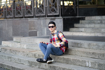 Fototapeta na wymiar Attractive teenage boy in checkered shirt, blue jeans, sunglasses and baseball cap sitting on a college campus on the stairs waiting for someone or something.Education technology lifestyle.Fashion guy