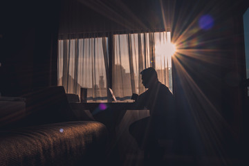 The silhouette of a man at work using a laptop in a modern room with a window background illuminated by slightly dazzling natural sun glare.