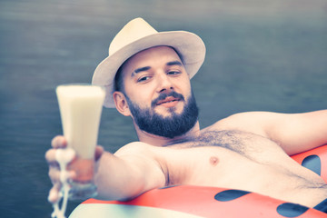 a man with a beard and a hat relaxes on a mattress with a drink. summer holiday concept