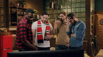 Four friends watching american football game, showing frustration. Group of young friends watching a football match, cheering and drinking beer, disappointed after they lost the match. Negative