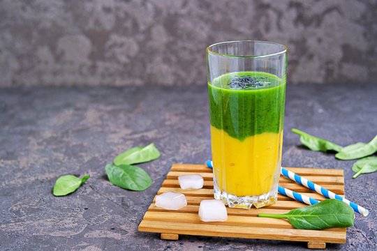 A healthy drink, a two-layer smoothie with fresh spinach and ripe mango in a tall glass glass on a dark concrete background. Drinks for detox and weight loss. Copyspace.