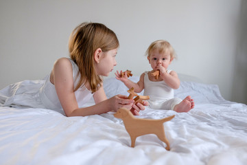 brother and sister in a white room are playing on a white bed. A boy in white clothes and a girl in a white dress play wooden safe and organic toys