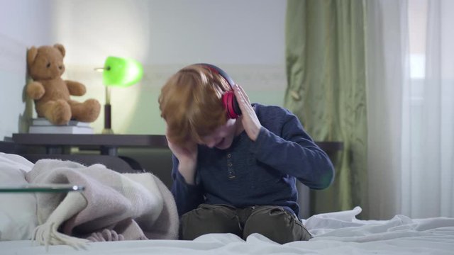 Little Caucasian boy with red hair putting on headphones and shaking head emotionally. Cute child listening to rock music indoors. Joy, hobby, lifestyle.