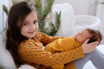 The older sister is sitting in an embrace with her newborn sister, who is swaddled,