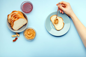 Top view,flat lay process of cooking breakfast, spreading bread, toast with peanut butter, creamy peanut paste by female hands on blue colored background. Served by peanut butter, peanuts in shell,jam