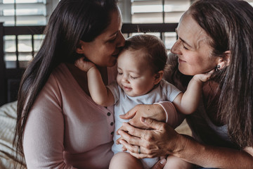 Happy baby girl holding cheeks of both smiling moms in front of window