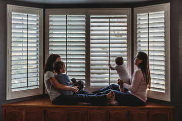 Same sex parents lounging with kids on window seat with tall shutters