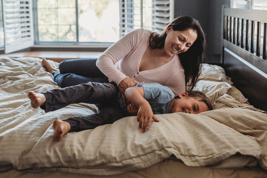 Laughing mom with long hair tickling 5 yr old son on bed near window