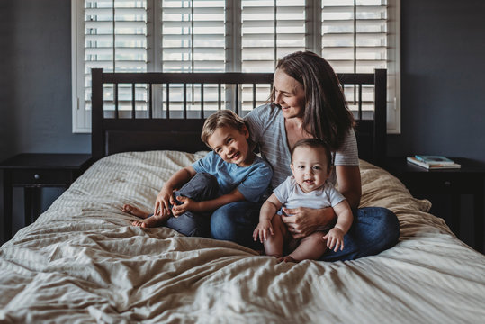 Laughing mom cuddling happy young son and baby on bed near window