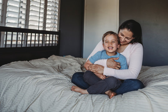 Happy mom holding smiling son on lap in bedroom near window