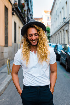 Stylish trendy long blonde haired guy smiling in the street with a hat