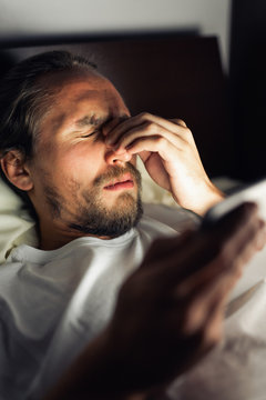 Bearded caucasian male with a headache in bed holding a tablet.