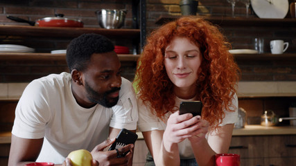 Portrait young african American man eat apple with curly red woman use their smart phone couple happy smile talk at home at kitchen girlfriend internet browsing technology together date slow motion