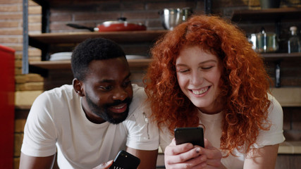 Close up african American man eat apple with curly red woman use their smart phone couple happy smile talk at home at kitchen girlfriend internet browsing technology together date shopping slow motion
